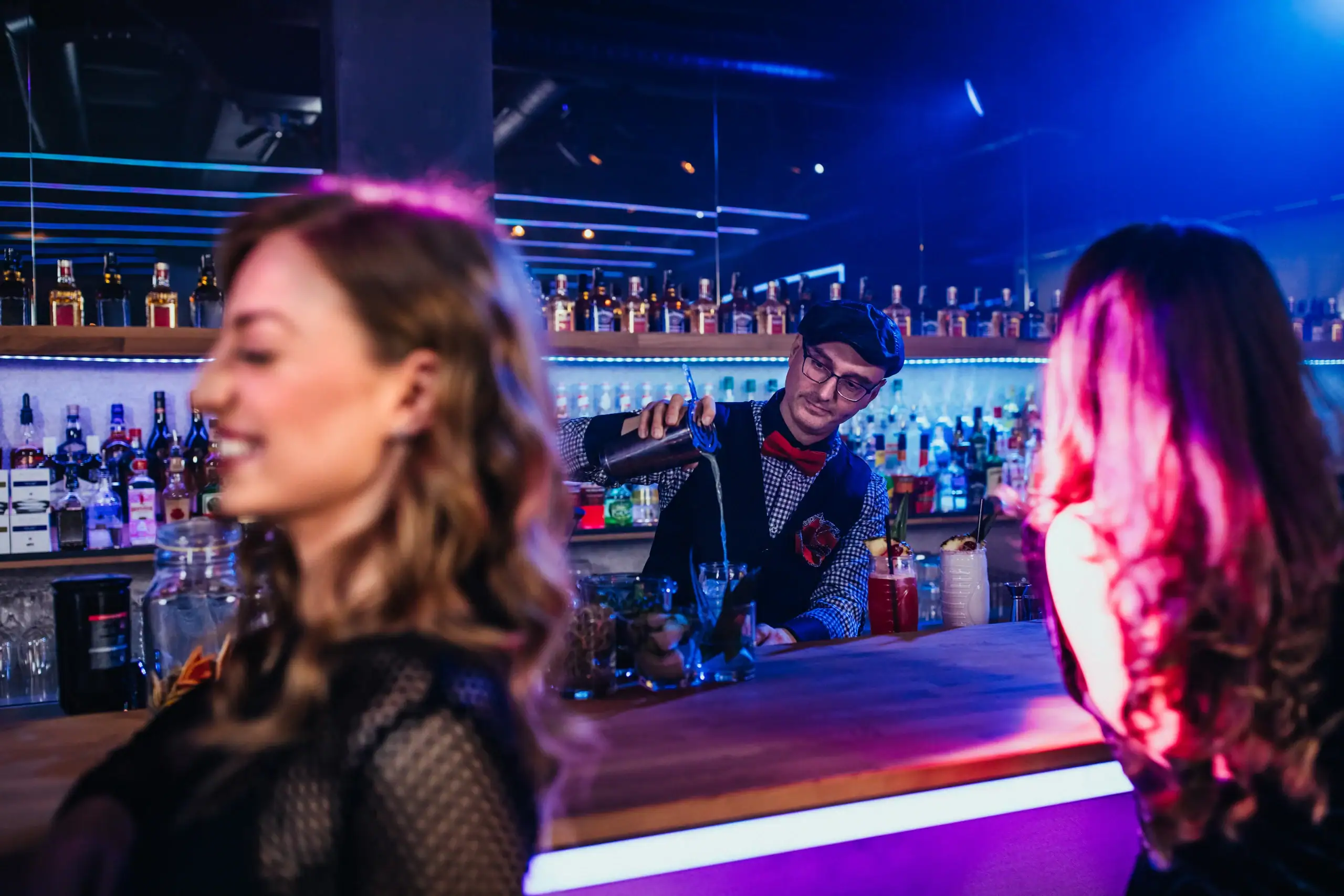 A bartender pours drinks behind a bar in a dimly lit nightclub, with two women in focus in the foreground, enjoying their evening. Bottles of alcohol are lined up on shelves in the background.