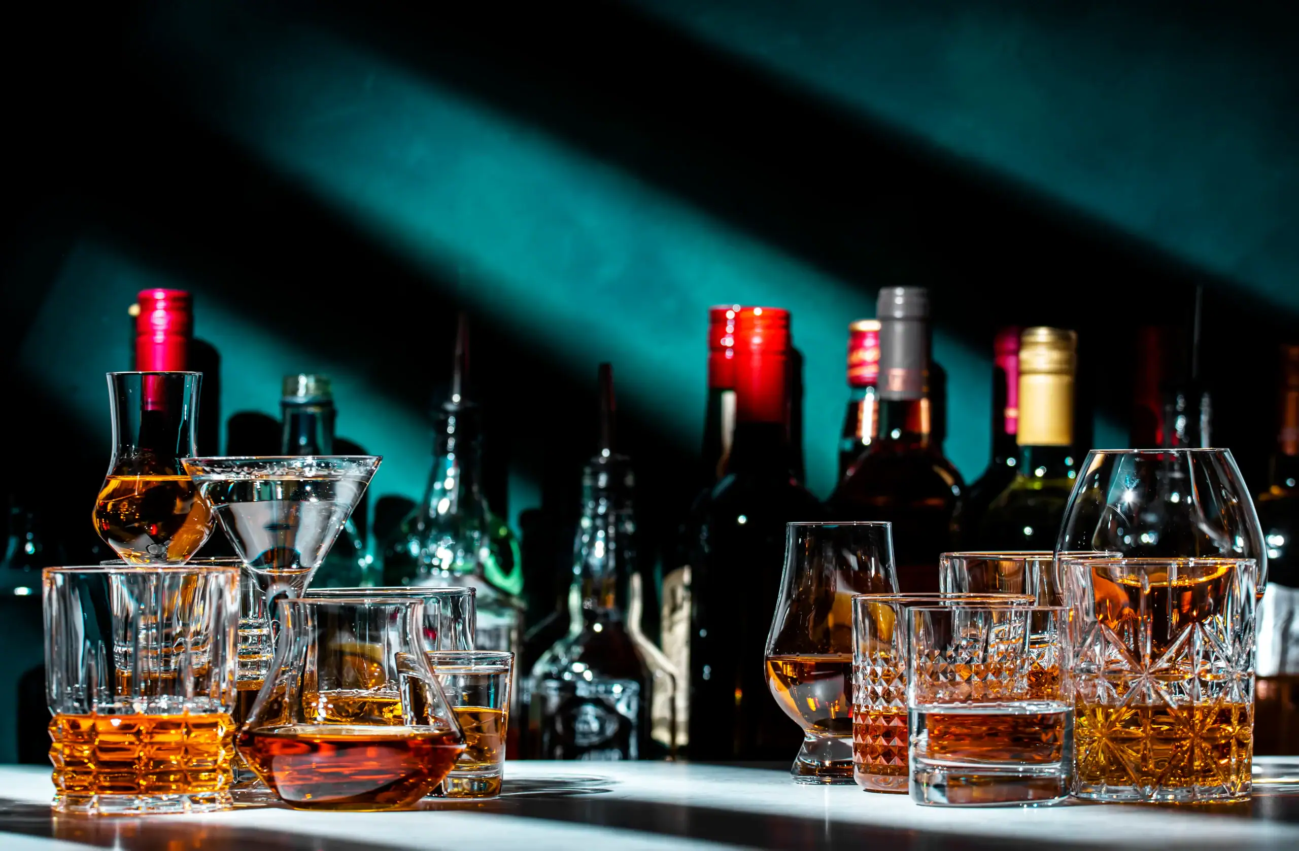A variety of whiskey and cocktail glasses filled with amber and clear drinks are arranged on a bar counter, with bottles of liquor and spirits blurred in the background under dramatic lighting.