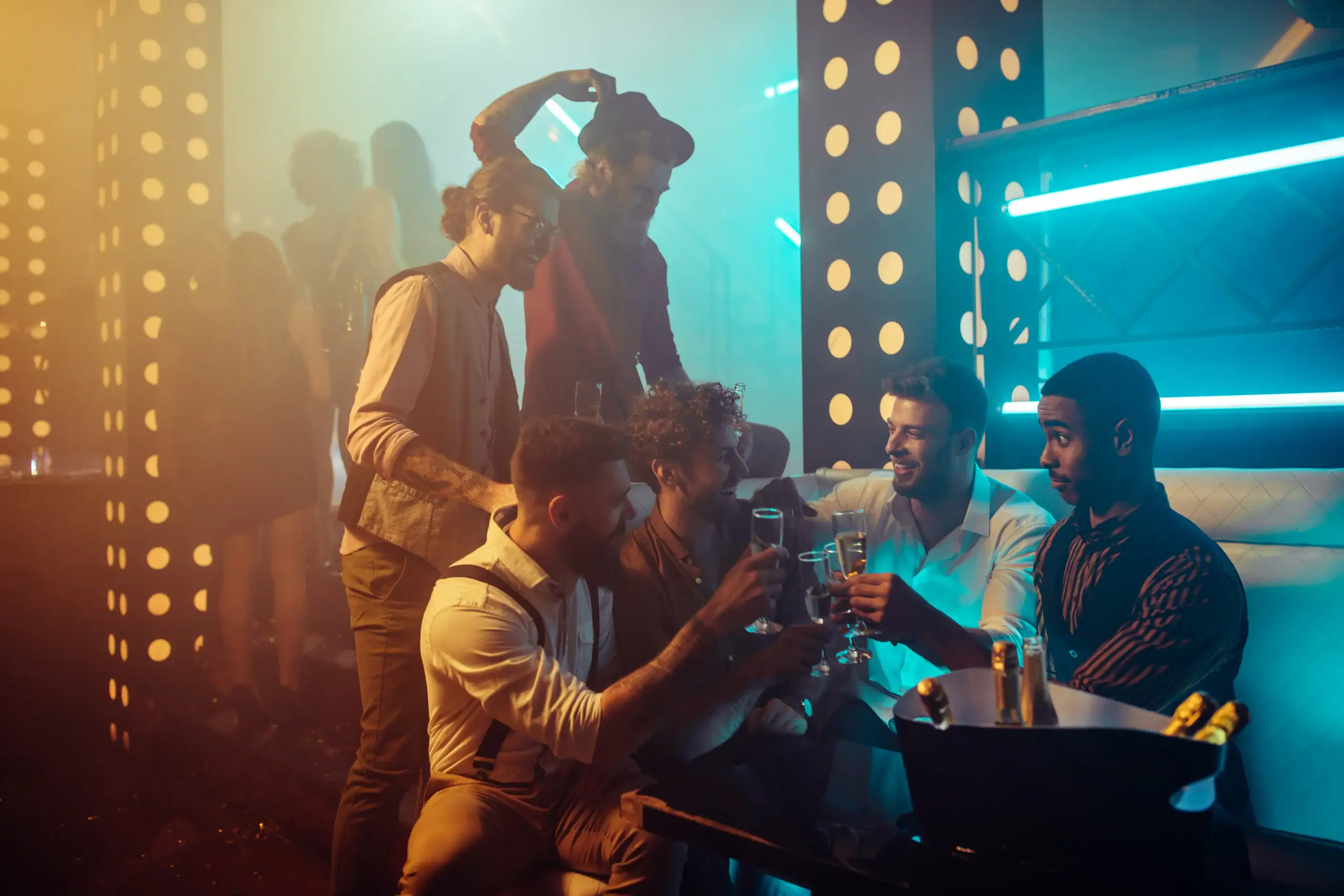 A group of young men in a club lounge clink glasses and celebrate, surrounded by neon lights and partygoers dancing in the background.