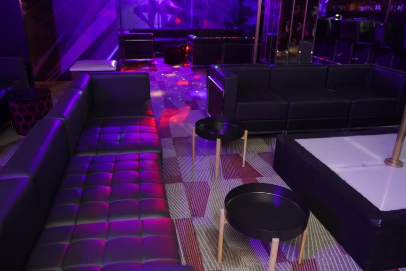 A lounge area with black leather sofas, small round black tables, and colorful lighting reflecting on the carpeted floor, creating a vibrant, club-like atmosphere.