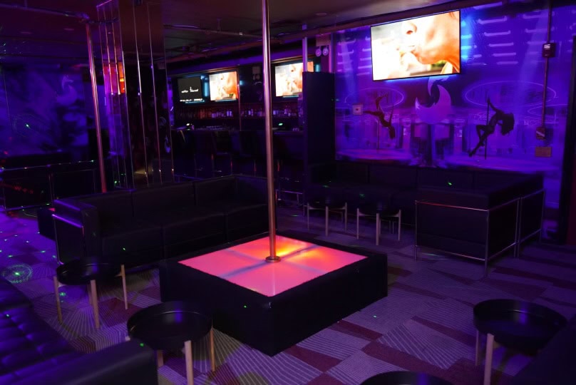 A dimly lit nightclub with purple lights, black couches, small round tables, a pole in the center, and TV screens on the wall showing videos. The atmosphere appears modern and stylish.