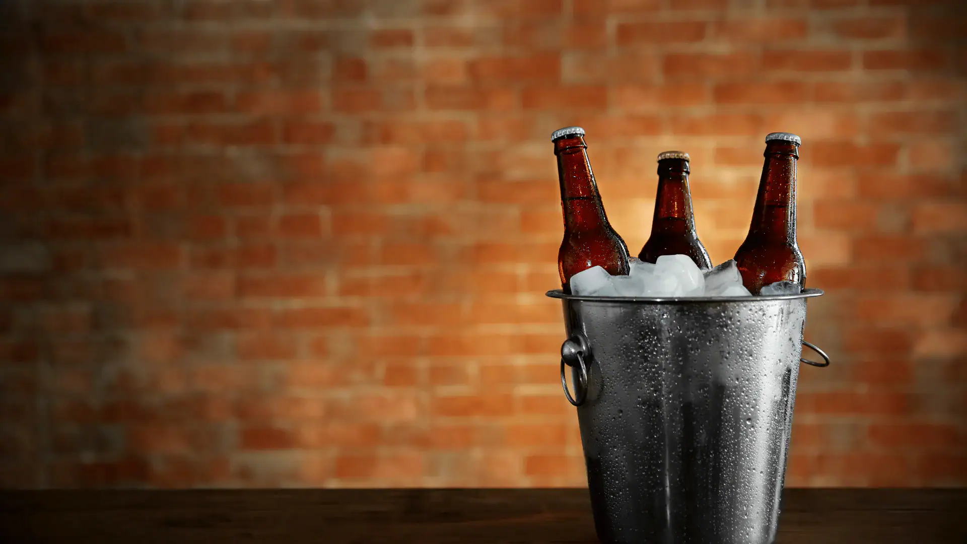 A metal bucket filled with ice holds three brown beer bottles. The bucket sits on a dark wooden surface, with a blurred red brick wall in the background.
