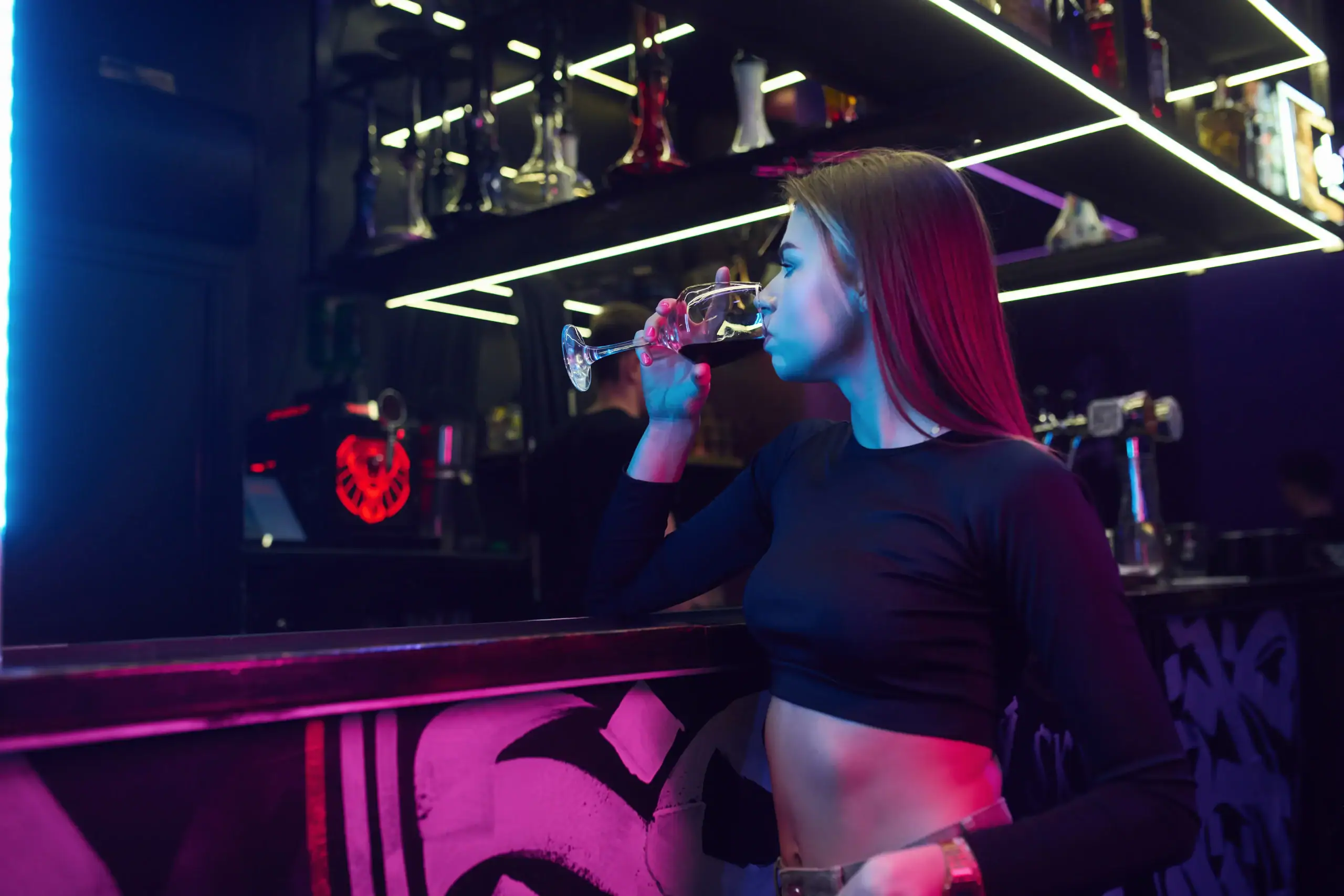 A young woman with long hair, wearing a black crop top, stands at a bar counter drinking from a wine glass under neon lights in a modern, stylish nightclub. Shelves with bottles and bar decor are visible in the background.